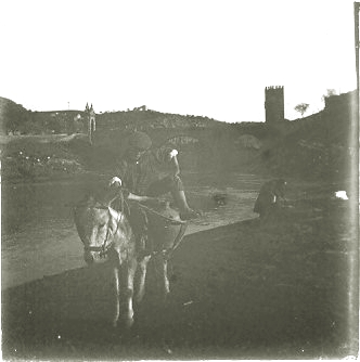 Fotografía que muestra en primer plano a un hombre sobre un burro que porta además dos cátaras de agua. Están a la orilla del río Tajo, en Toledo. Al fondo, el puente de Alcántara. Detrás del hombre, una mujer se inclina sobre un objeto no identificado.