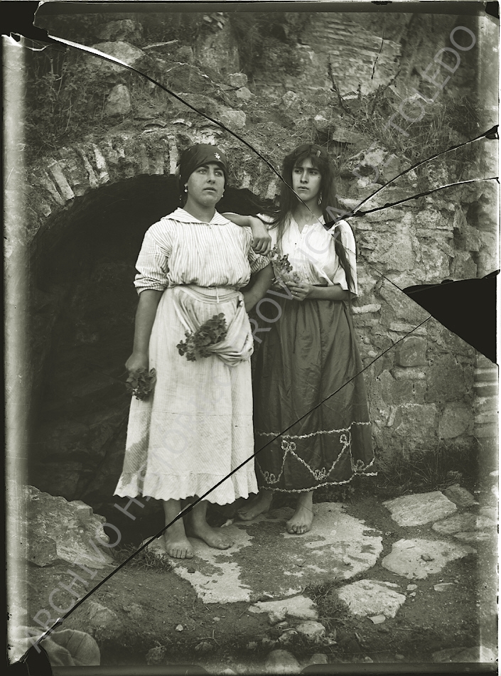 Dos chicas jóvenes de apariencia gitana, descalzas y con pequeños ramilletes de flores en sus manos, a la puerta de una especie de cueva.